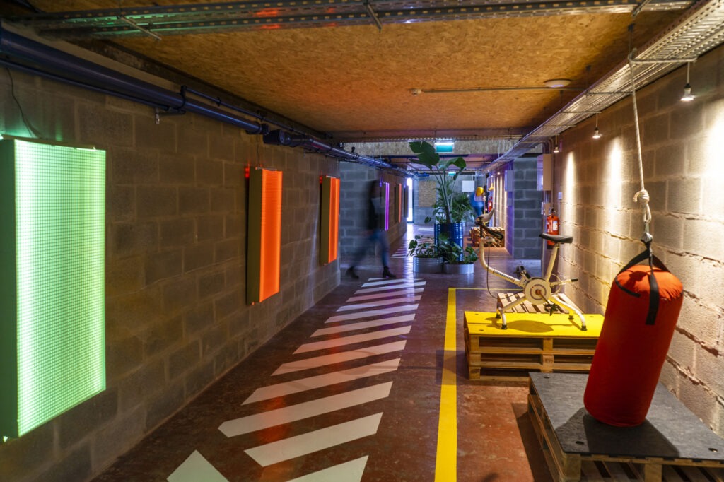 Corredor de entrada do SITIO Alto de São João, com painéis luminosos coloridos e sinalização branca no chão